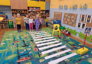 Dzieci w sali przedszkolnej przechodzą po pasach z białego papieru jako przejście dla pieszych a nauczyciel pokazuje koło w kolorze zielonym.
