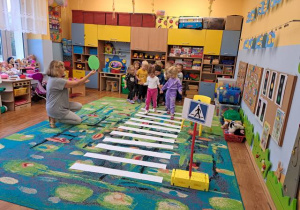 Dzieci w sali przedszkolnej przechodzą po pasach z białego papieru jako przejście dla pieszych a nauczyciel pokazuje koło w kolorze zielonym.
