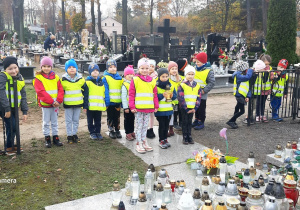 Grupa dzieci stoją naprzeciwko pomnika zbiorowej mogiły na cmentarzu.