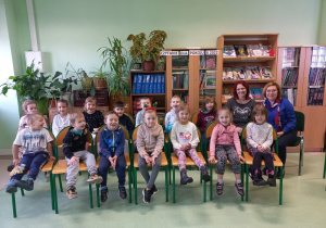 Grupa dzieci siedzi na krzesłach w bibliotece.
