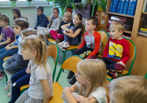 Grupa dzieci siedzących na krzesłach słucha czytanej książki.