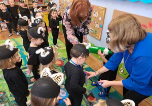 Dzieci w strojach kotków są pasowane na Przedszkolaka dużą zieloną kredką przez Panią Dyrektor