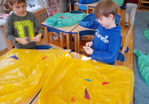 Dzieci na dywanie ozdabiają worki na śmieci kolorowymi wzorami z folii samoprzylepnej.