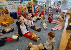 Dzieci huśtają misia siedząc w siadzie prostym.