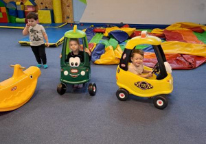 Dzieci w sali zabaw – dwoje w samochodach zabawkowych i jedno stoi obok