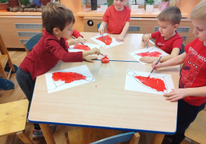 Dzieci siedzą przy stolikach i malują farbami szablon czapki Mikołaja.