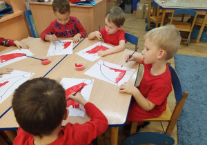 Dzieci siedzą przy stolikach i malują farbami szablon czapki Mikołaja.