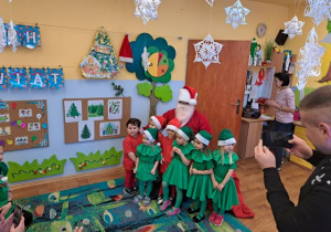 Trzech chłopców w czerwonych strojach i cztery dziewczynki w zielonych strojach pozują do zdjęcia ze Świętym Mikołajem