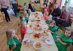 Grupa dzieci siedzi przy stole ze słodkim poczęstunkiem, a obok nich rodzice i rodzeństwo