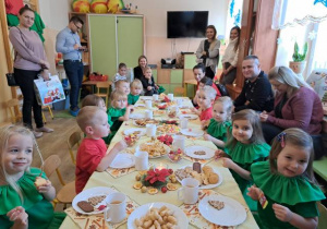 Grupa dzieci siedzi przy stole ze słodkim poczęstunkiem, a obok nich rodzice i rodzeństwo