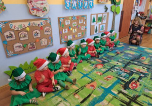 Grupa dzieci -dziewczynki w zielonych strojach, jako choineczki i chłopcy w czerwonych strojach, jako Mikołaje, występują przed rodzicami.