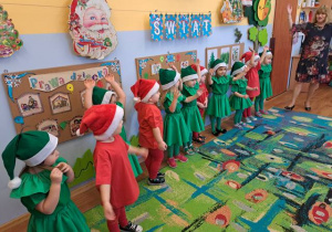 Grupa dzieci -dziewczynki w zielonych strojach, jako choineczki i chłopcy w czerwonych strojach, jako Mikołaje, występują przed rodzicami.