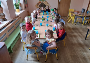 Grupa dzieci w strojach karnawałowych siedzi przy stoliku ze słodkim poczęstunkiem