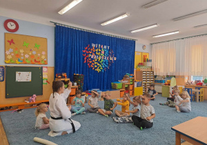 Dzieci siedzą w kole na dywanie wokoło trenerki karate.