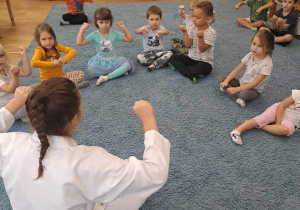 Dzieci siedzą w kole na dywanie wokoło trenerki karate.