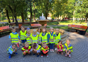 Grupa dzieci w kamizelkach odblaskowych pozuje do zdjęcia a w tle pomnik Rafała Bratoszewskiego założyciela miasta Aleksandrów Łódzki.
