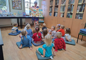 Grupa dzieci siedzi w bibliotece na poduszkach na podłodze a przed nimi pani bibliotekarka pokazuje największą książkę znajdującą w bibliotece.