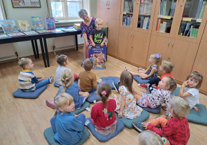 Grupa dzieci siedzi w bibliotece na poduszkach na podłodze a przed nimi pani bibliotekarka wraz z chłopcem pokazuje największą książkę znajdującą w bibliotece.