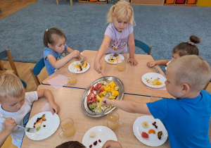 Grupa dzieci przy stolikach wykonuje sałatkę owocową.