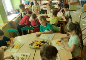 Dzieci siedzą przy stolikach i przy użyciu plastikowych noży kroją owoce na deskach do krojenia