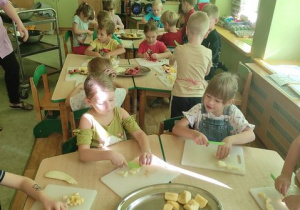 Dzieci siedzą przy stolikach i kroją owoce na deskach do krojenia