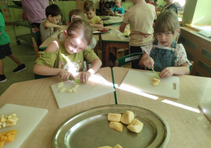 Dwie dziewczynki kroją owoce na sałatkę, w tle inne dzieci przy stoliku