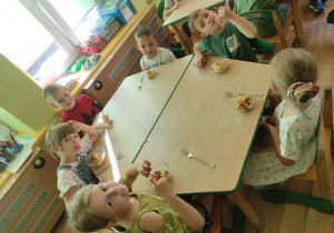Dzieci siedzą przy stolikach, przed sobą mają sałatkę owocową w salaterkach