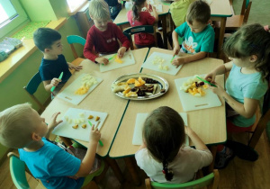 Sześcioro dzieci siedzi przy stoliku i kroi samodzielnie wybrane przez siebie owoce na sałatkę
