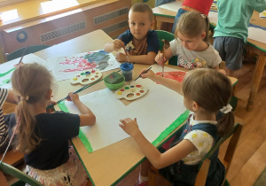 Dzieci siedząc przy stoliku malują farbami na białym kartonie