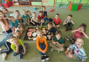 Dzieci pozują na dywanie z plakatem, na którym naklejone są pokolorowane przez nie owoce