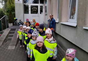 Grupa dzieci w żółtych kamizelkach (ustawionych w parach) stoi przed przedszkolem.