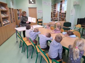 Grupa dzieci siedzi przy stolikach w bibliotece a pani bibliotekarka czyta książkę.