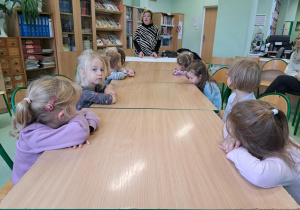 Grupa dzieci siedzi przy stolikach w bibliotece i słucha wypowiedzi pani bibliotekarki