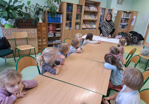 Grupa dzieci siedzi przy stolikach w bibliotece i słucha wypowiedzi pani bibliotekarki