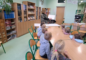 Grupa dzieci siedzi przy stolikach w bibliotece a pani bibliotekarka czyta książkę