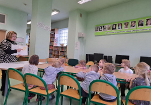 Grupa dzieci siedzi przy stolikach w bibliotece a pani bibliotekarka czyta książkę