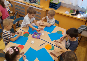 Dzieci siedząc przy stolikach tworzą jesienne drzewa z kół origami