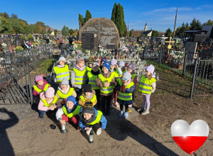 Grupa dzieci stoi na cmentarzu przy zbiorowej mogile żołnierzy, którzy zginęli podczas II Wojny Światowej. Niektóre dzieci trzymają znicze.