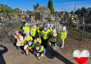 Grupa dzieci stoi na cmentarzu przy zbiorowej mogile żołnierzy, którzy zginęli podczas II Wojny Światowej, w tym część dzieci trzyma znicze.