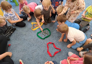 Dzieci siedzą w kole na dywanie, grupują elementy w 3 szarfach