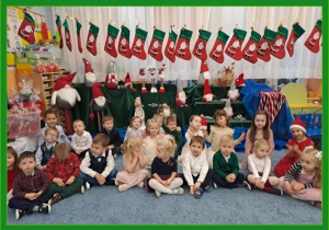 Dzieci w świątecznych strojach stoją siedzą pod girlandą ze świątecznych skarpet.