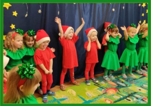 Dzieci w świątecznych przebraniach stoją na świątecznym tle, jedno z nich wesoło unosi ręce do góry.