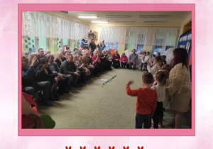 Zdjęcie w różowej ramce przedstawiające grupę dzieci ustawionych w rzędzie i machających do dziadków na widowni.