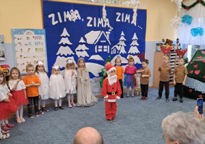 Dzieci w różnych strojach podczas występu dla babć i dziadków, na pierwszym planie chłopiec w stroju św. Mikołaja.