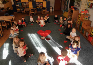 Dzieci siedząc na dywanie trzymają czerowne serduszka z papieru.