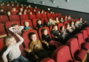 Dzieci siedzące na czerwonych fotelach w sali kinowej.