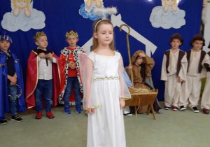 Dziewczynka-aniołek recytuje tekst przedstawienia, w tle dzieci występujące w Jasełkach