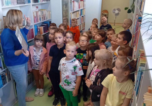 Dzieci zwiedzają bibliotekę i słuchają pani bibliotekarki, która opowiada o swojej pracy.