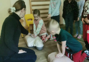 Prowadząca zajęcia obserwuje dwoje dzieci ćwiczące usiskanie klatki piersiowej przy resuscytacji (na fantomach).