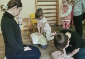 Prowadząca zajęcia obserwuje dwoje dzieci ćwiczące usiskanie klatki piersiowej przy resuscytacji (na fantomach).